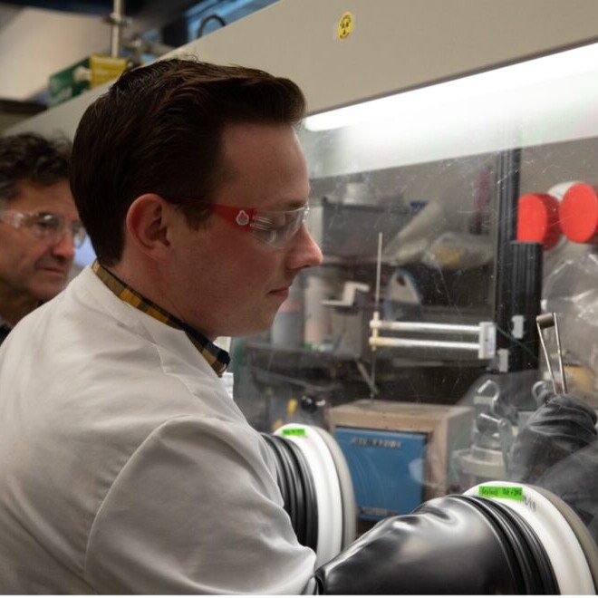 Jarom Chamberlain in lab coat and lab glasses reaching into glass display as Professor Michael Simpson in gray flannel looks on