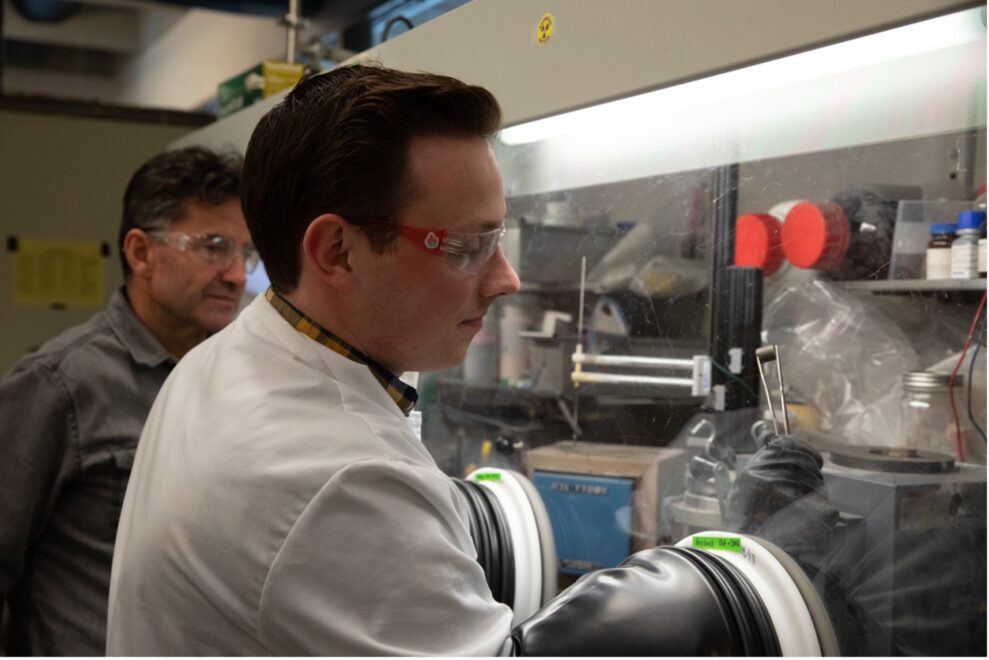 Jarom Chamberlain in lab coat and lab glasses reaching into glass display as Professor Michael Simpson in gray flannel looks on