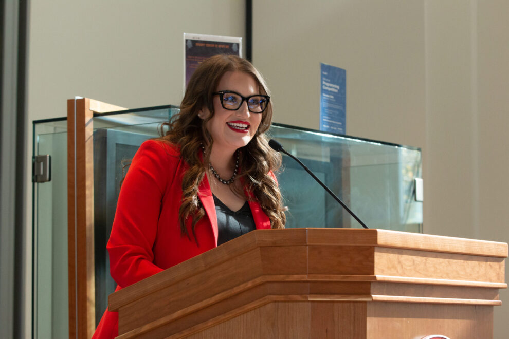 Heather Kahlert with black glasses wearing a red suit jacket and standing at a podium