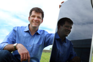 A man with a blue short crouching next to a black solar panel. University of Utah associate professor Mike Scarpulla