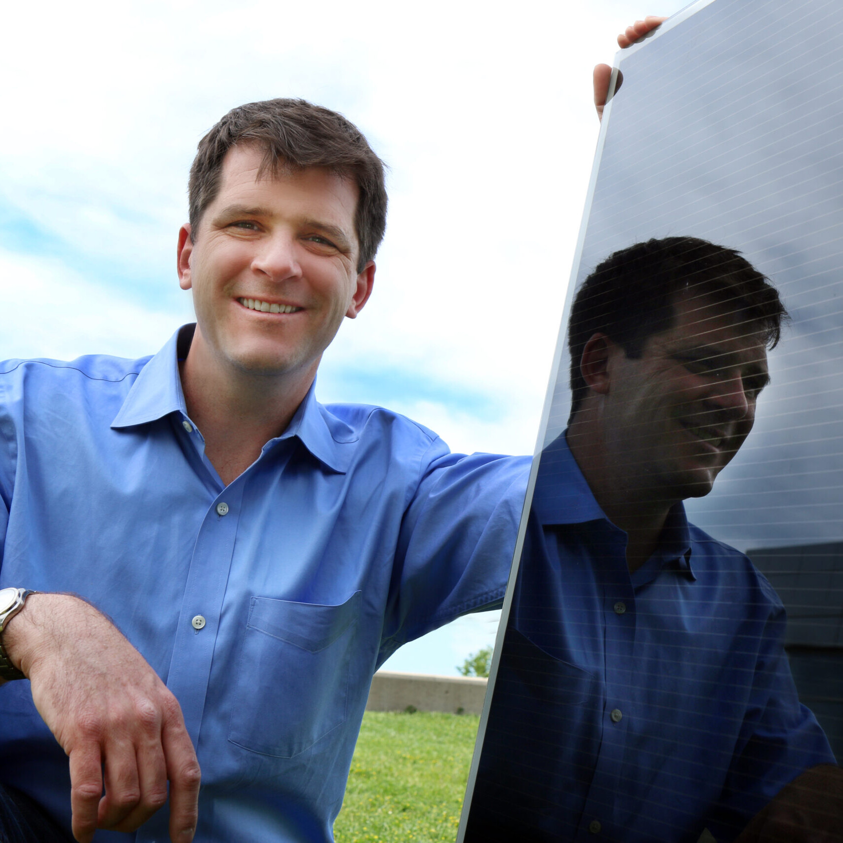 A man with a blue short crouching next to a black solar panel. University of Utah associate professor Mike Scarpulla