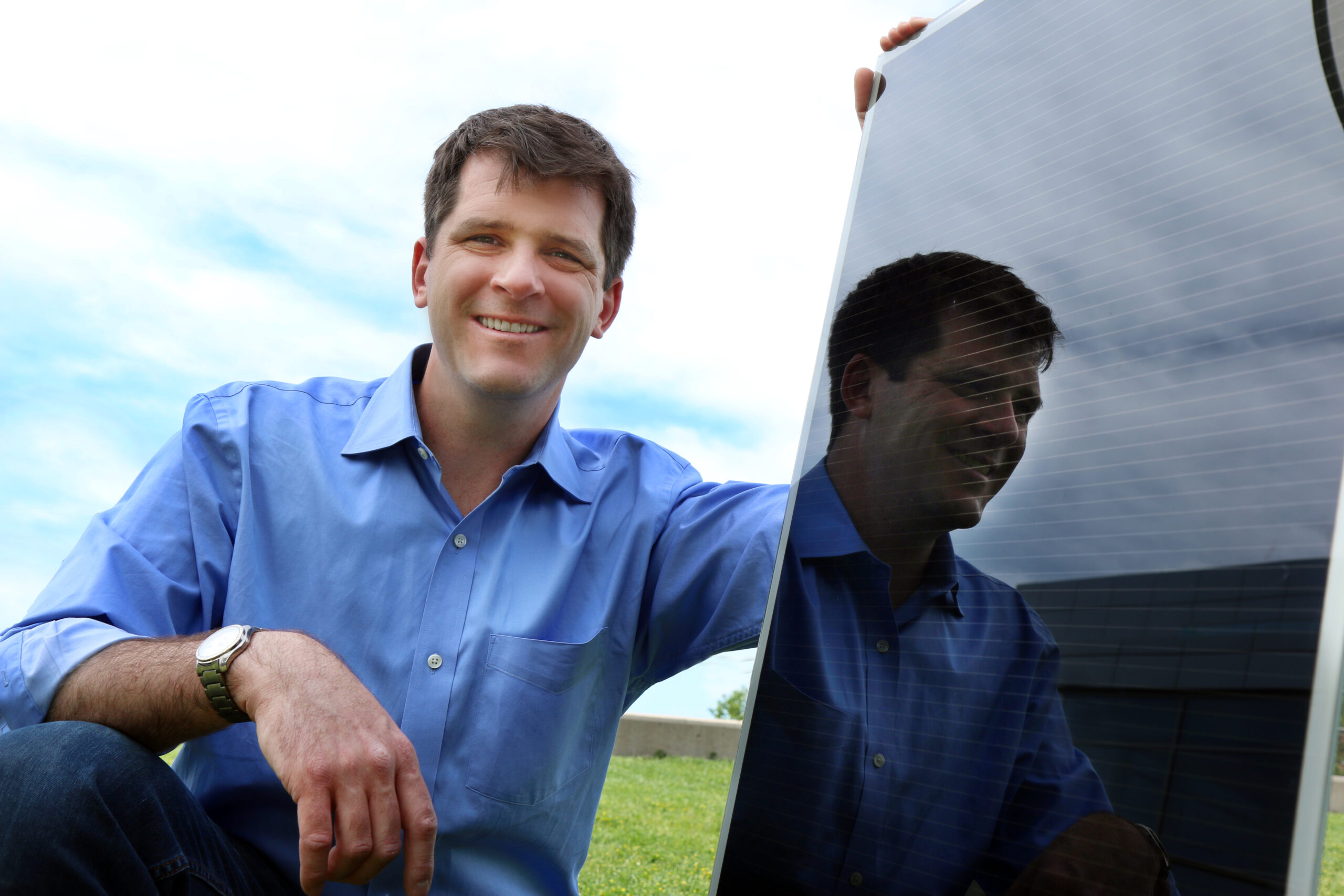 A man with a blue short crouching next to a black solar panel. University of Utah associate professor Mike Scarpulla