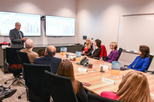 A man in a dark suit gives a presentation to a group of people in a conference room.