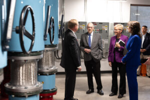 Two men and two women standing next to large blue pipes in a geothermal pump room.