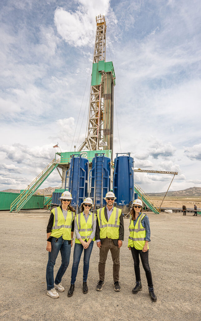 Four people in safety vests stand in front of a towering geothermal well drill