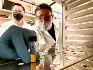 A researcher in masks and gloves holds a vial full of a yellow powder.