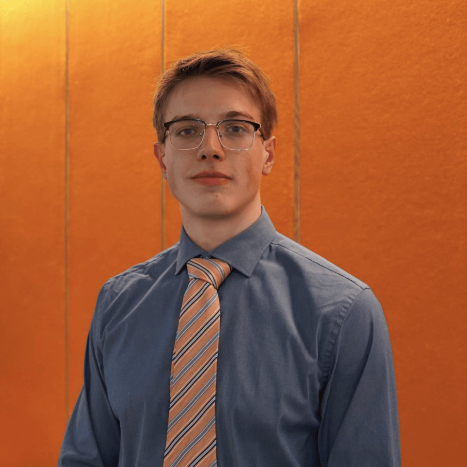headshot of Maxim Balitskiy. He is posed against an orange background in a steel grey shirt and striped tie