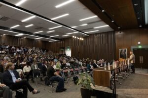 A speaker addresses a large crowd in a wood-paneled lecture hall.