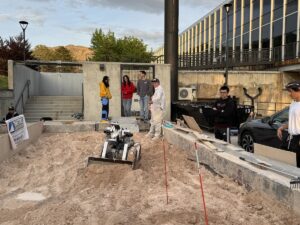 A bulldozer shaped robot drives in what looks like an oversized sandbox as students look on.