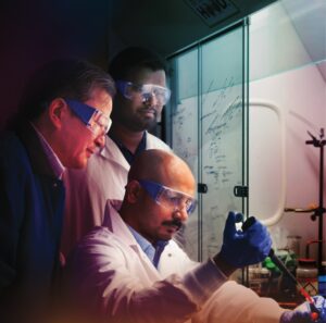 Three researchers in front of a chemical hood, transferring a glowing liquid into a vial